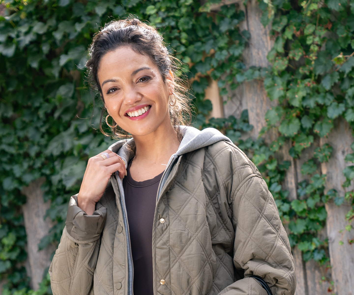 A woman wearing an olive quilted jacket over a dark top, standing outdoors against a wall covered in green plants.