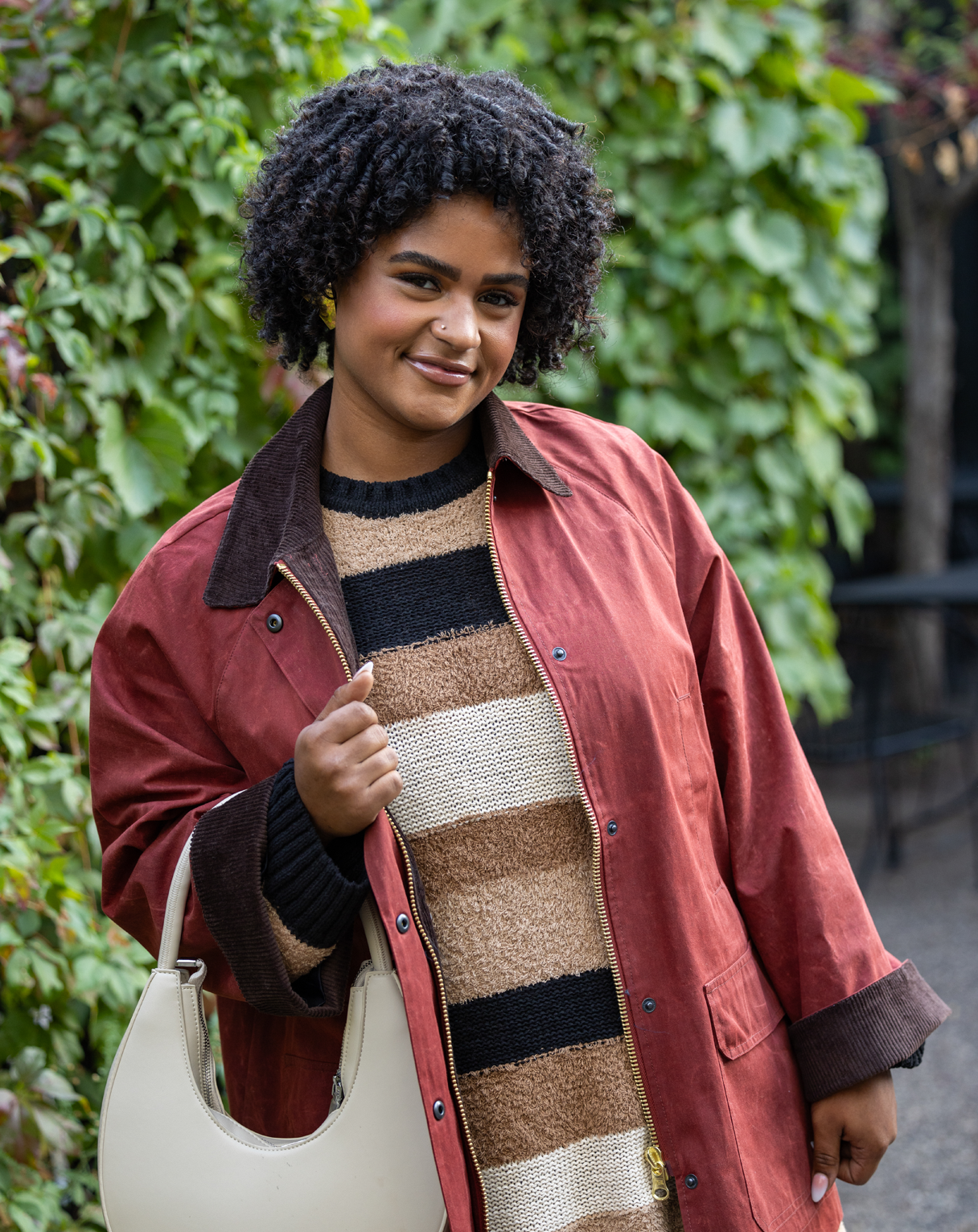 A woman wearing a red coat over a striped sweater, holding a cream handbag, standing near greenery.