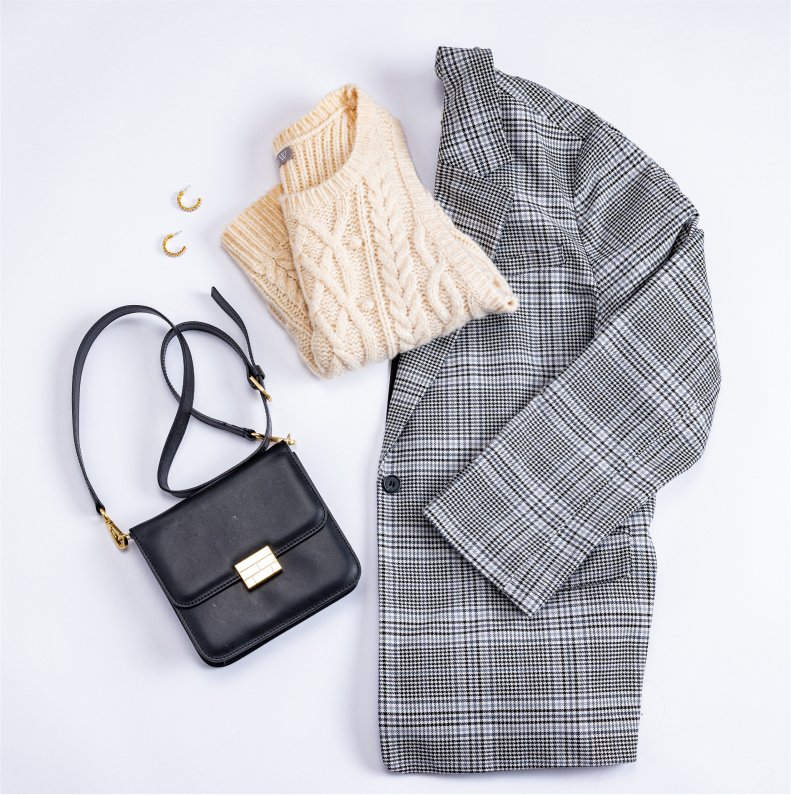 A neatly folded plaid blazer, white knit sweater, a black purse, and earrings all load out on a flat white surface