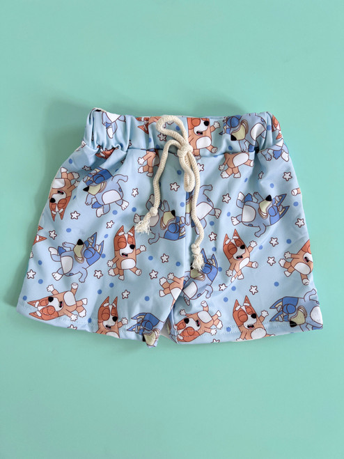 Baby and little boys' swim trunks in light blue featuring a fun character print and star background. The trunks have an elastic waistband with a faux cream drawstring detail.
