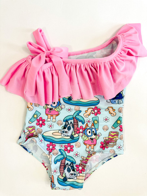 Little girl's one-piece swimsuit with a vibrant character print in blue and a pink ruffle detail at the shoulders adorned with a bow.