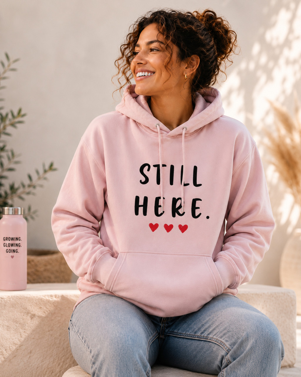 Woman wearing a light pink hoodie featuring the empowering message “STILL HERE.” and three red hearts while seated indoors in a bright, cozy setting. Inspirational apparel symbolizing resilience, strength, perseverance, hope, and overcoming life's challenges.
