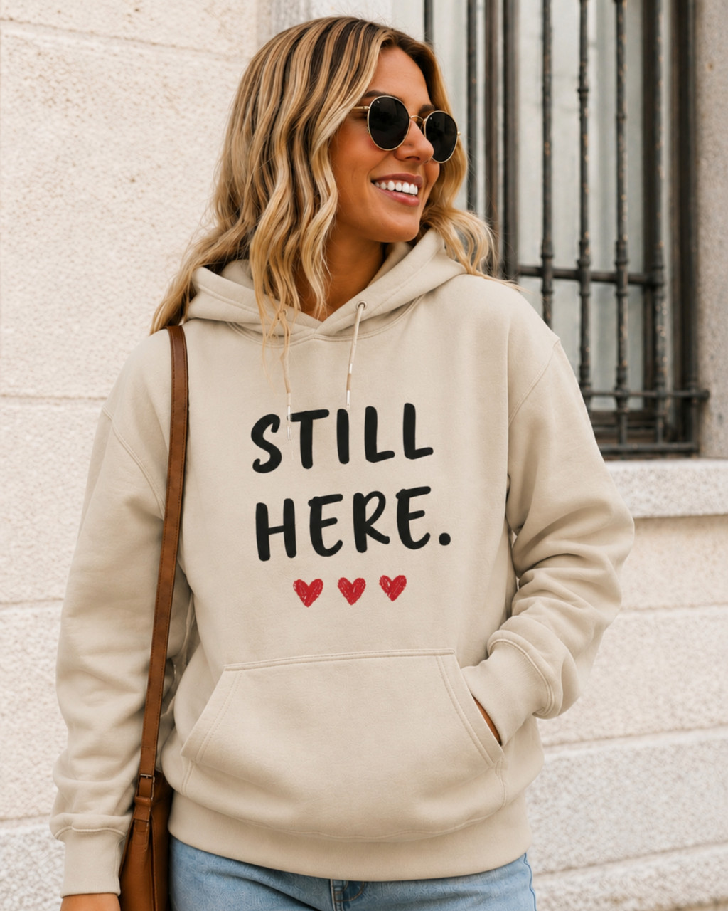 Woman modeling a sand-colored hoodie displaying the inspirational message “STILL HERE.” with three red hearts. Motivational unisex sweatshirt ideal for encouragement gifts, resilience awareness, emotional support, and everyday casual comfort.