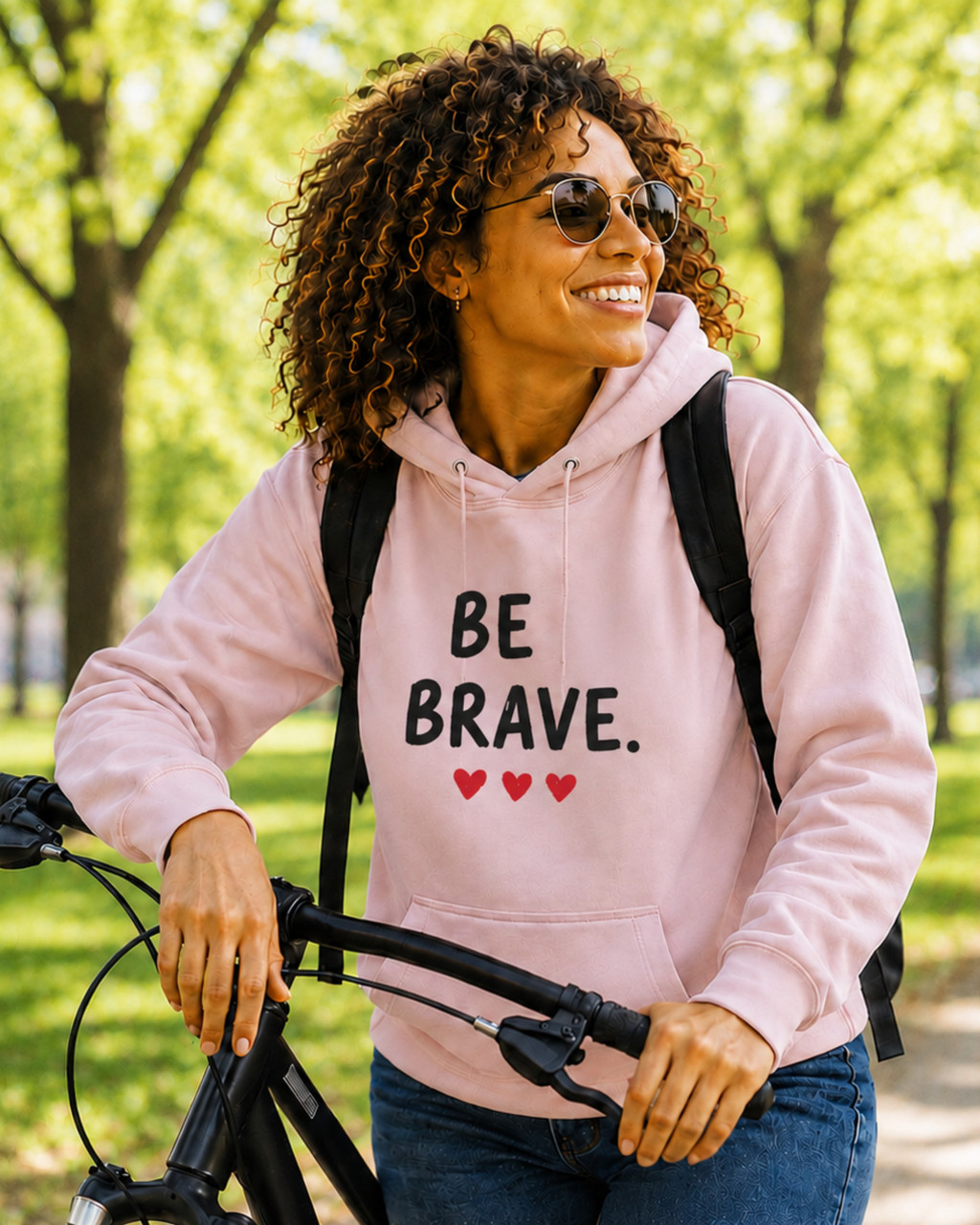 Woman modeling a blush pink motivational hoodie featuring the “BE BRAVE” graphic and three red hearts while outdoors with a bicycle. Inspirational apparel designed to encourage confidence, resilience, and self-belief.