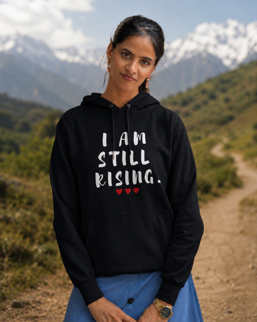 Woman wearing a black hoodie with the inspirational message “I AM STILL RISING” and red heart accents, standing on a scenic mountain trail with snow-capped peaks in the background. Motivational women's apparel for resilience, strength, personal growth, and empowerment.