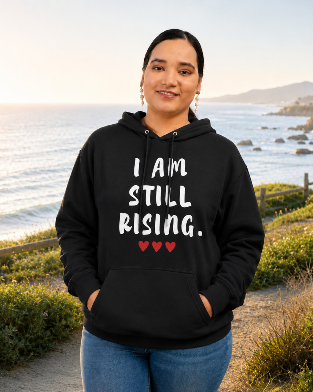 Women's black motivational hoodie featuring “I AM STILL RISING” graphic and red hearts, photographed on a coastal overlook. Inspirational apparel designed to celebrate resilience, perseverance, self-belief, empowerment, and overcoming challenges.