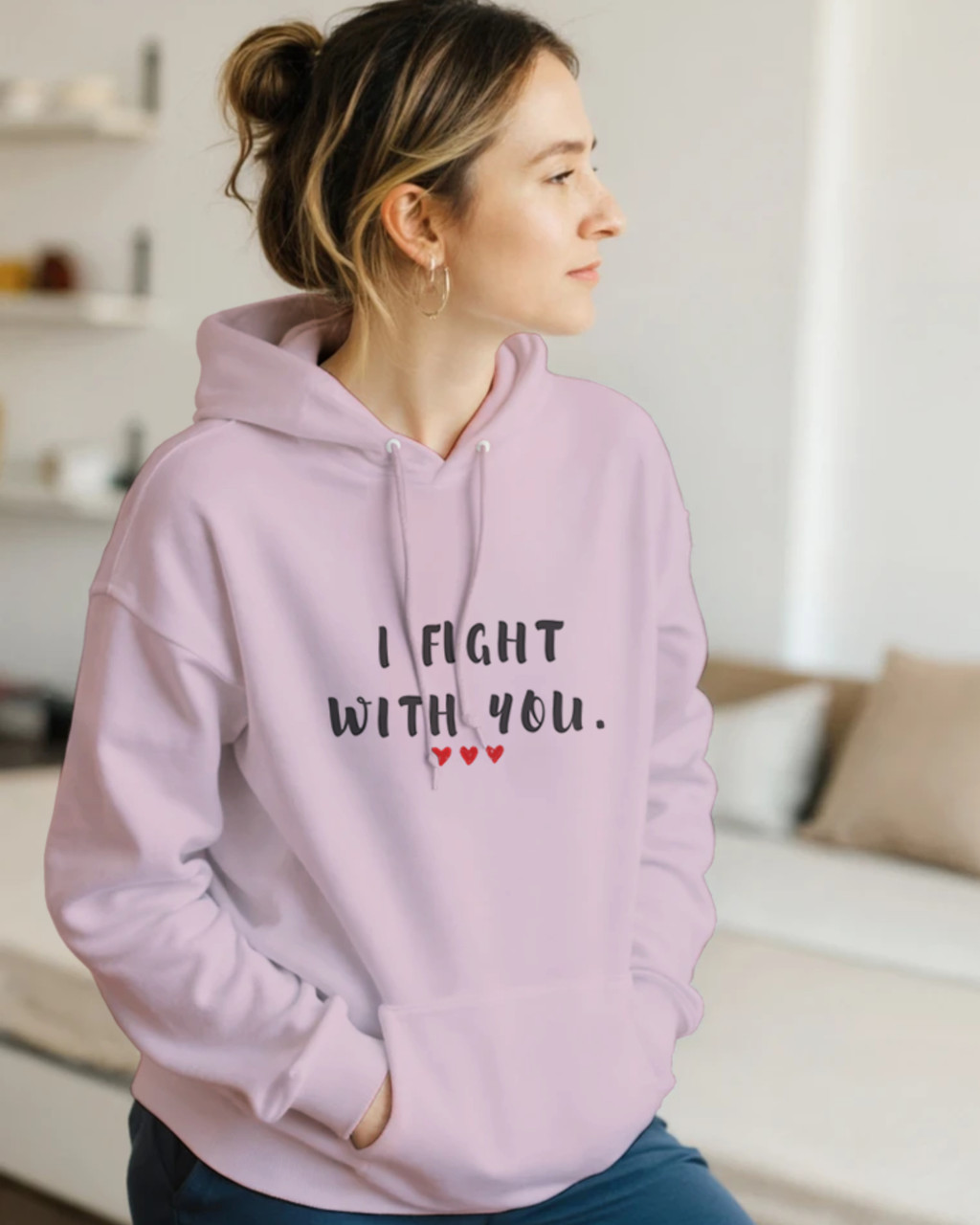 Woman wearing a light pink pullover hoodie featuring the supportive phrase “I Fight With You” in bold black lettering with three red hearts beneath. The hoodie has a relaxed fit, drawstring hood, and front kangaroo pocket. The uplifting design symbolizes strength, solidarity, encouragement, and standing together through life’s challenges.