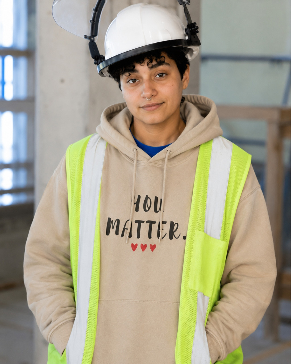 Construction worker wearing a sand-colored pullover hoodie featuring the uplifting phrase “You Matter” in distressed black lettering with three red hearts beneath. The hoodie is worn under a high-visibility safety vest and hard hat at a job site, symbolizing encouragement, self-worth, resilience, and support. The hoodie includes a drawstring hood, front kangaroo pocket, and relaxed unisex fit.