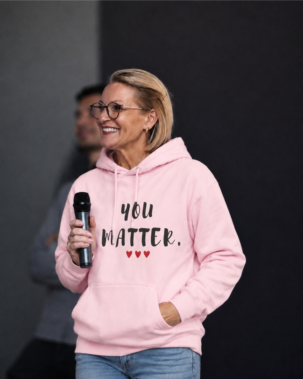 Woman wearing a light pink pullover hoodie featuring the uplifting phrase “You Matter” in distressed black lettering with three red hearts beneath. She is holding a microphone while speaking at an event, conveying confidence, encouragement, and positivity. The hoodie includes a drawstring hood, front kangaroo pocket, and relaxed fit.