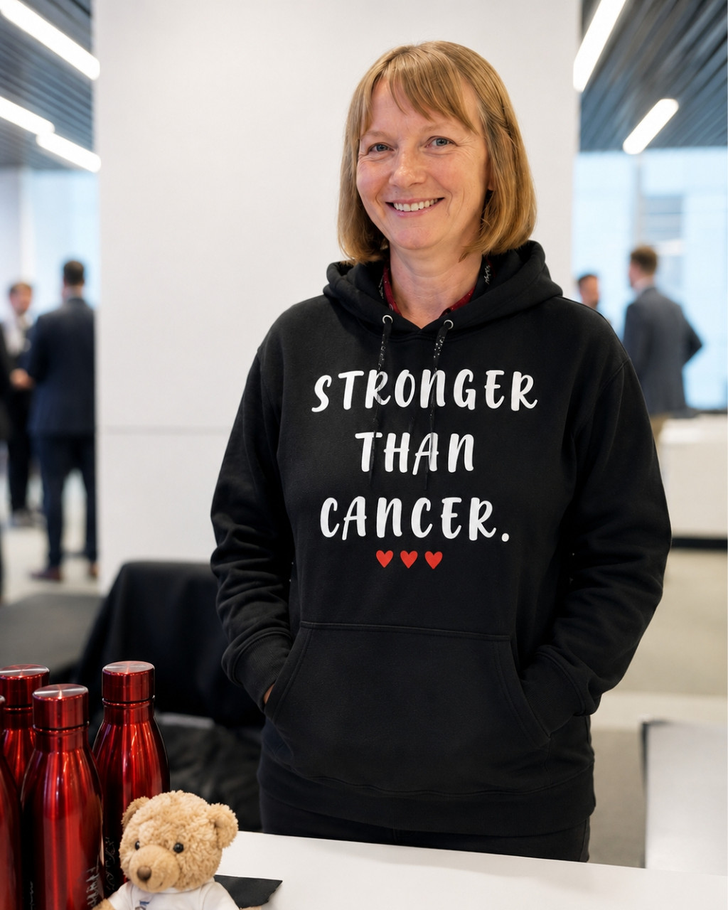 Smiling woman wearing a black hoodie with the inspirational message “STRONGER THAN CANCER.” printed in white lettering and three small red hearts underneath. She is standing indoors at a modern event or conference venue, with people blurred in the background. Several red metallic water bottles and a small teddy bear sit on the table in front of her. The hoodie design is clearly visible and serves as the focal point of the image, conveying strength, hope, and cancer awareness.