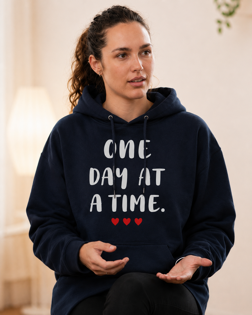 Woman seated indoors wearing a navy blue hoodie with the inspirational message “ONE DAY AT A TIME.” printed in large white lettering across the front, accented by three small red hearts beneath the text. She appears engaged in conversation or a support group discussion, gesturing with her hands while looking attentively to the side. The softly lit interior and calm expression create a warm, encouraging atmosphere. The design promotes hope, resilience, and taking life’s challenges one step at a time.