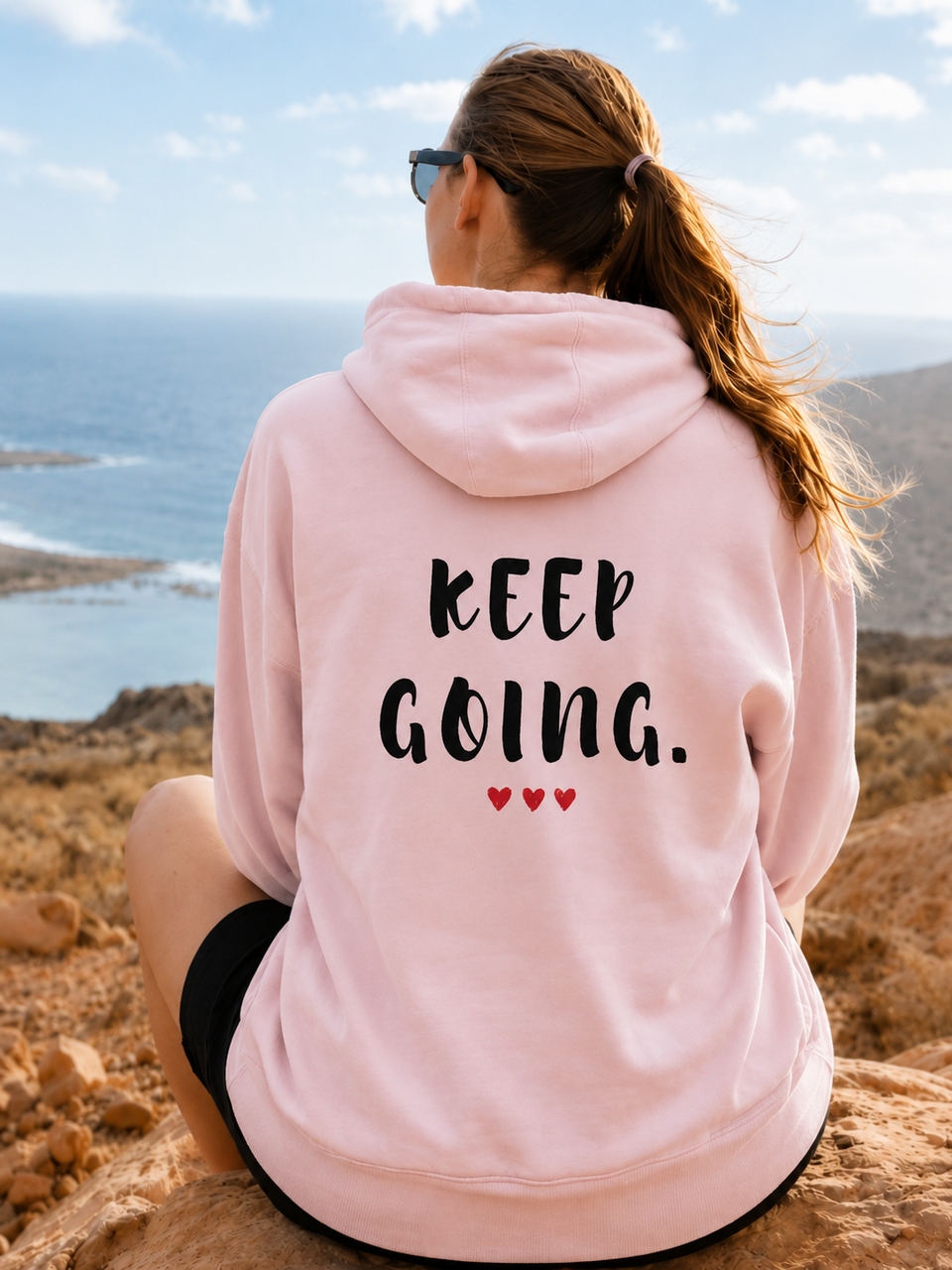 Rear view of a woman sitting on a rocky coastal overlook wearing a light pink hoodie with the inspirational message “KEEP GOING.” printed in bold black distressed lettering across the back. Three small red hearts appear beneath the text. She faces the ocean and distant shoreline under a bright sky, with her hair tied back in a ponytail. The scenic landscape and motivational design create a sense of perseverance, hope, resilience, and moving forward despite challenges.