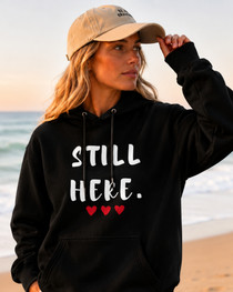 Female model wearing a black hoodie with the motivational phrase “STILL HERE.” and red heart accents. Meaningful sweatshirt promoting courage, determination, mental wellness, self-belief, and personal growth in a coastal setting.
