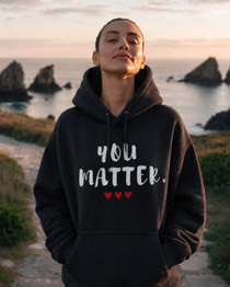 Woman standing on a scenic coastal path at sunset wearing a black pullover hoodie featuring the uplifting phrase “You Matter” in bold white lettering with three red hearts beneath. The hoodie has a relaxed fit, drawstring hood, and front kangaroo pocket. The inspirational design promotes self-worth, encouragement, hope, and positivity.