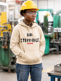 Person wearing a sand-colored pullover hoodie with the motivational message “UNSTOPPABLE.” printed in distressed black lettering and accented by three small red hearts beneath the text. They are standing in an industrial workshop while wearing a yellow hard hat and safety glasses, with their hands tucked into the hoodie’s kangaroo pocket. Machinery and manufacturing equipment are visible in the softly blurred background. The image conveys determination, resilience, confidence, and perseverance while showcasing the hoodie design in a hardworking, professional environment.