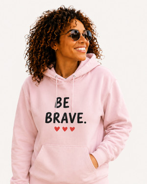 Woman modeling a blush pink motivational hoodie featuring the “BE BRAVE” graphic and three red hearts while outdoors with a bicycle. Inspirational apparel designed to encourage confidence, resilience, and self-belief.