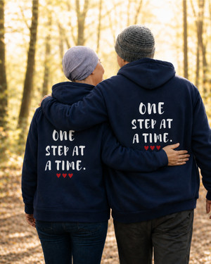 Two people wearing matching navy blue hoodies with the quote “ONE STEP AT A TIME” and red hearts, walking arm in arm on a forest trail, symbolizing hope, support, resilience, friendship, and personal growth.
