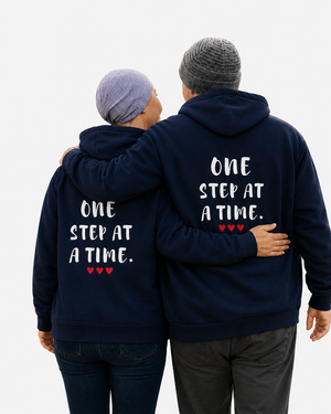 Two people wearing matching navy blue hoodies with the quote “ONE STEP AT A TIME” and red hearts, walking arm in arm on a forest trail, symbolizing hope, support, resilience, friendship, and personal growth.