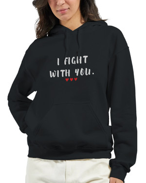 Woman walking down stairs with coffee cup wearing a black pullover hoodie featuring the inspirational phrase “I Fight With You” in bold white lettering with three red hearts beneath. The hoodie has a drawstring hood, front kangaroo pocket, and relaxed fit, making it a meaningful symbol of support, strength, and solidarity.