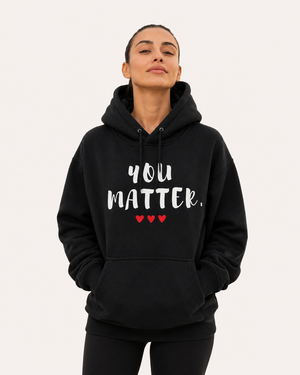 Woman standing on a scenic coastal path at sunset wearing a black pullover hoodie featuring the uplifting phrase “You Matter” in bold white lettering with three red hearts beneath. The hoodie has a relaxed fit, drawstring hood, and front kangaroo pocket. The inspirational design promotes self-worth, encouragement, hope, and positivity.