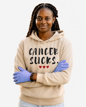 Young woman wearing a sand-colored pullover hoodie featuring the phrase “Cancer Sucks.” in distressed black lettering with three red hearts beneath. She stands outdoors in a park while participating in a community clean-up event. The hoodie combines a message of cancer awareness with themes of compassion, resilience, support, and positive action.