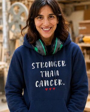 Smiling woman wearing a navy blue hoodie with the inspirational message “STRONGER THAN CANCER.” printed in distressed white lettering and three small red hearts underneath. She is standing indoors in a workshop or makerspace environment, with tools and equipment softly blurred in the background. Protective earmuffs rest around her neck, adding a practical, hardworking feel to the scene. The hoodie design is prominently displayed, conveying strength, resilience, hope, and support for those affected by cancer.