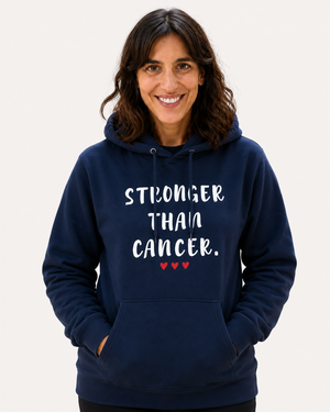 Smiling woman wearing a navy blue hoodie with the inspirational message “STRONGER THAN CANCER.” printed in distressed white lettering and three small red hearts underneath. She is standing indoors in a workshop or makerspace environment, with tools and equipment softly blurred in the background. Protective earmuffs rest around her neck, adding a practical, hardworking feel to the scene. The hoodie design is prominently displayed, conveying strength, resilience, hope, and support for those affected by cancer.