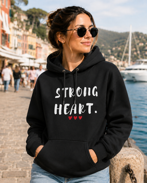 Woman wearing a black hoodie with the empowering message “STRONG HEART.” printed in distressed white lettering and accented by three small red hearts beneath the text. She is standing along a scenic waterfront promenade, wearing sunglasses and smiling as she looks into the distance. Colorful buildings, people strolling, boats, and a marina create a vibrant coastal backdrop. The image conveys strength, resilience, confidence, and hope while showcasing the hoodie design in a stylish outdoor setting.