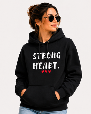 Woman wearing a black hoodie with the empowering message “STRONG HEART.” printed in distressed white lettering and accented by three small red hearts beneath the text. She is standing along a scenic waterfront promenade, wearing sunglasses and smiling as she looks into the distance. Colorful buildings, people strolling, boats, and a marina create a vibrant coastal backdrop. The image conveys strength, resilience, confidence, and hope while showcasing the hoodie design in a stylish outdoor setting.