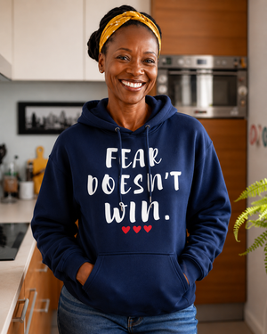 Woman wearing a navy blue hoodie with the motivational quote “FEAR DOESN’T WIN” and three red hearts, smiling indoors in a modern kitchen, symbolizing courage, resilience, hope, confidence, and overcoming life’s challenges.