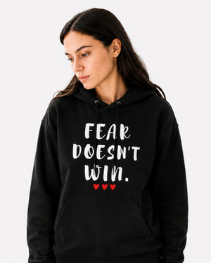 Woman wearing a navy blue hoodie with the motivational quote “FEAR DOESN’T WIN” and three red hearts, smiling indoors in a modern kitchen, symbolizing courage, resilience, hope, confidence, and overcoming life’s challenges.