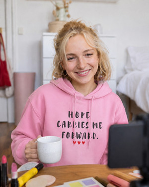 Woman wearing a pink hoodie with the inspirational quote “Hope Carries Me Forward” in distressed black lettering and red hearts, smiling while working at a desk in a cozy home setting.