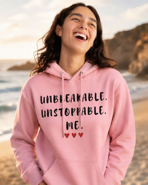 Woman wearing a pink hoodie featuring the empowering quote “Unbreakable. Unstoppable. Me.” with heart accents, smiling on a beach, inspirational apparel celebrating confidence, resilience, strength, and self-belief.