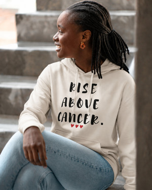 Woman wearing a beige hoodie featuring the inspirational quote “Rise Above Cancer” with red heart accents, seated on outdoor steps, motivational apparel promoting hope, courage, resilience, strength, and cancer awareness.