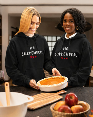 Two women wearing matching black hoodies featuring the inspirational quote “No Surrender” with red heart accents, smiling while baking together in a kitchen, motivational apparel celebrating resilience, determination, strength, and perseverance.