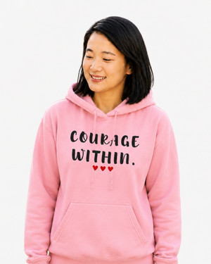 Woman wearing a pink Gildan 18500 hoodie featuring the “Courage Within” inspirational graphic, smiling while using a tablet outdoors in a sunlit garden setting.