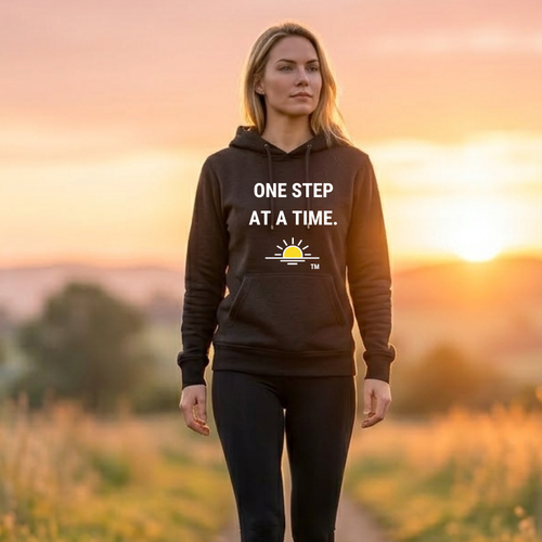 A person is standing outdoors on a path during sunset. Warm golden light fills the background, softly blurring the surrounding landscape. The person is wearing a black hooded sweatshirt with bold white text across the chest that reads “ONE STEP AT A TIME.” The serene lighting and simple composition draw attention to the motivational message on the hoodie.