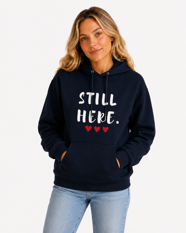Female model seated on a sofa wearing a navy blue hoodie with the motivational phrase “STILL HERE.” and red heart accents. Meaningful sweatshirt promoting courage, mental wellness, determination, and personal growth.