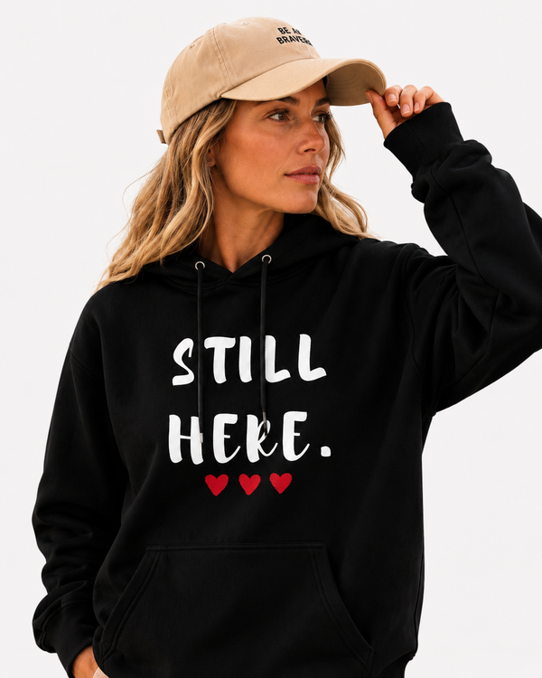 Female model wearing a black hoodie with the motivational phrase “STILL HERE.” and red heart accents. Meaningful sweatshirt promoting courage, determination, mental wellness, self-belief, and personal growth in a coastal setting.