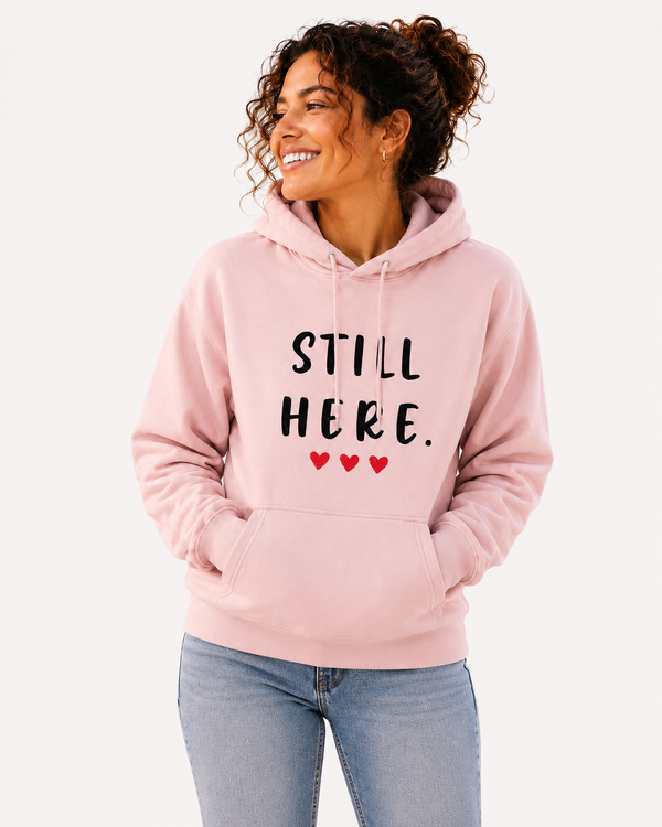 Woman wearing a light pink hoodie featuring the empowering message “STILL HERE.” and three red hearts while seated indoors in a bright, cozy setting. Inspirational apparel symbolizing resilience, strength, perseverance, hope, and overcoming life's challenges.