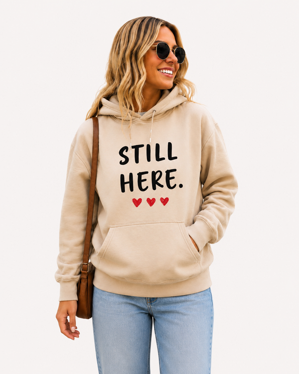 Woman modeling a sand-colored hoodie displaying the inspirational message “STILL HERE.” with three red hearts. Motivational unisex sweatshirt ideal for encouragement gifts, resilience awareness, emotional support, and everyday casual comfort.