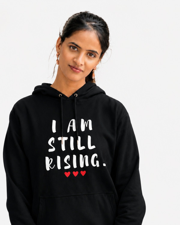 Woman wearing a black hoodie with the inspirational message “I AM STILL RISING” and red heart accents, standing on a scenic mountain trail with snow-capped peaks in the background. Motivational women's apparel for resilience, strength, personal growth, and empowerment.