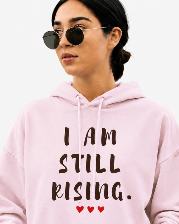 Women's light pink motivational hoodie with “I AM STILL RISING” graphic and red heart details, photographed in a modern city environment. Inspirational apparel designed to celebrate strength, empowerment, recovery, determination, and positive mindset.