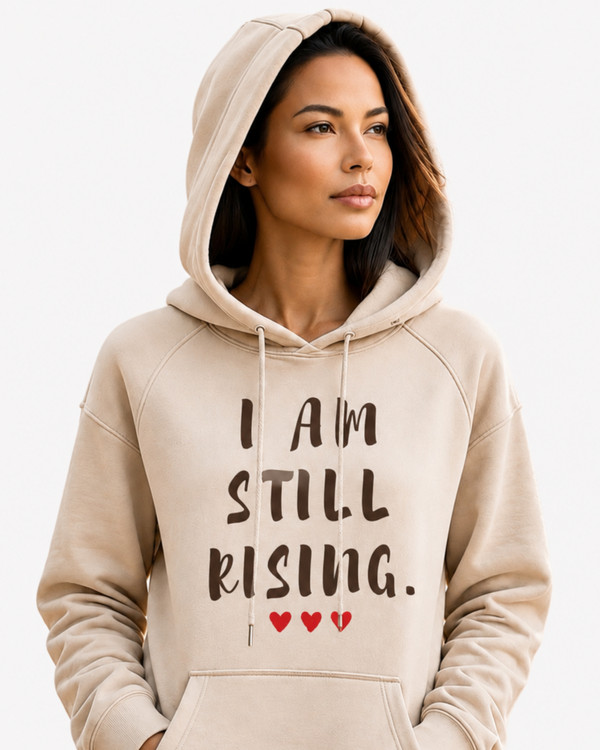 Woman wearing a sand-colored inspirational hoodie featuring the message “I AM STILL RISING” with red heart accents, standing on a sunlit forest trail. Motivational women's apparel promoting resilience, personal growth, confidence, healing, and empowerment.