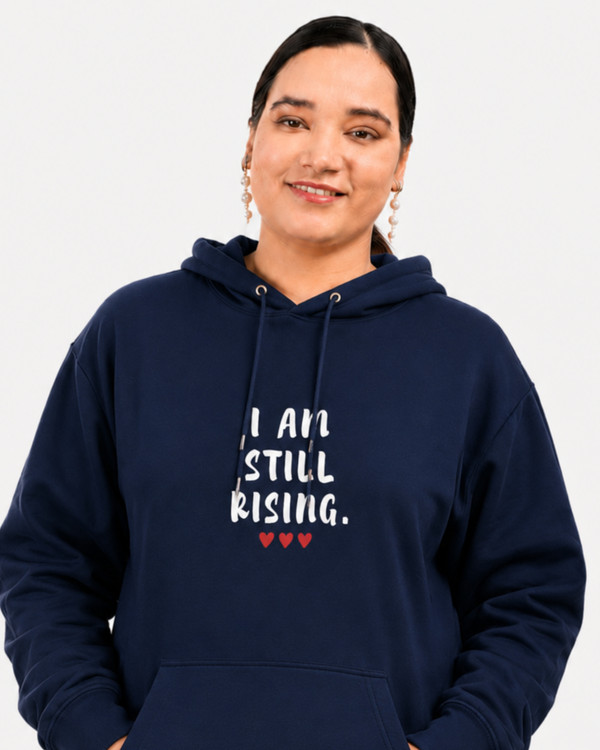 Women's black motivational hoodie featuring “I AM STILL RISING” graphic and red hearts, photographed on a coastal overlook. Inspirational apparel designed to celebrate resilience, perseverance, self-belief, empowerment, and overcoming challenges.
