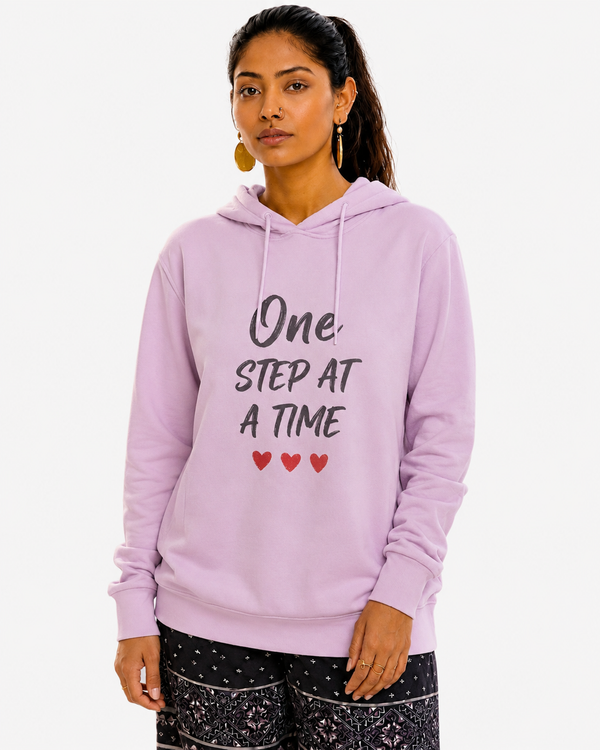 Woman wearing a pink hoodie with the motivational quote “One Step At A Time” and three red hearts, standing on a beach at sunset, symbolizing hope, perseverance, resilience, and personal growth.