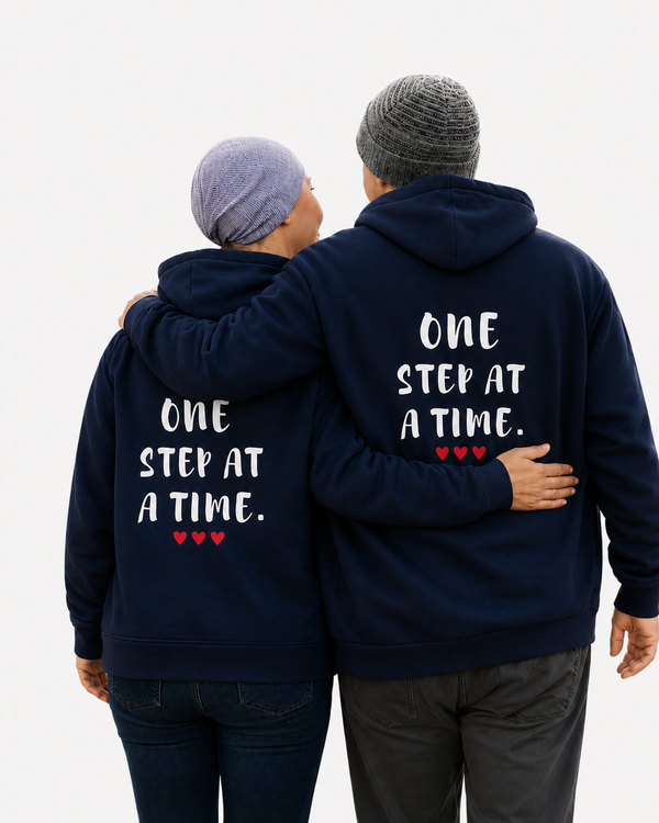 Two people wearing matching navy blue hoodies with the quote “ONE STEP AT A TIME” and red hearts, walking arm in arm on a forest trail, symbolizing hope, support, resilience, friendship, and personal growth.