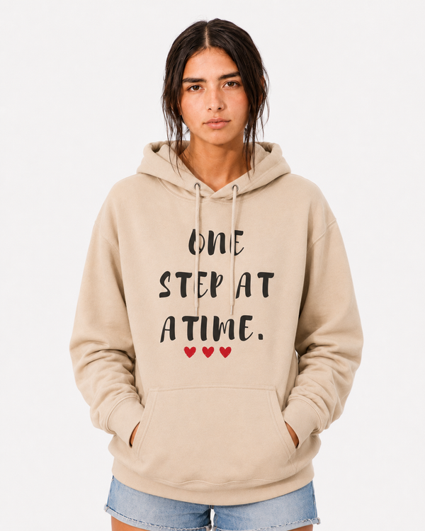 Woman wearing a sand-colored hoodie with the inspirational quote “ONE STEP AT A TIME” and three red hearts, standing outdoors in a rural field, symbolizing resilience, hope, perseverance, healing, and personal growth.