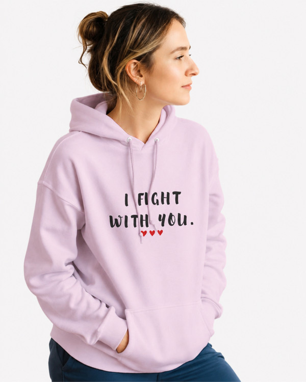 Woman wearing a light pink pullover hoodie featuring the supportive phrase “I Fight With You” in bold black lettering with three red hearts beneath. The hoodie has a relaxed fit, drawstring hood, and front kangaroo pocket. The uplifting design symbolizes strength, solidarity, encouragement, and standing together through life’s challenges.