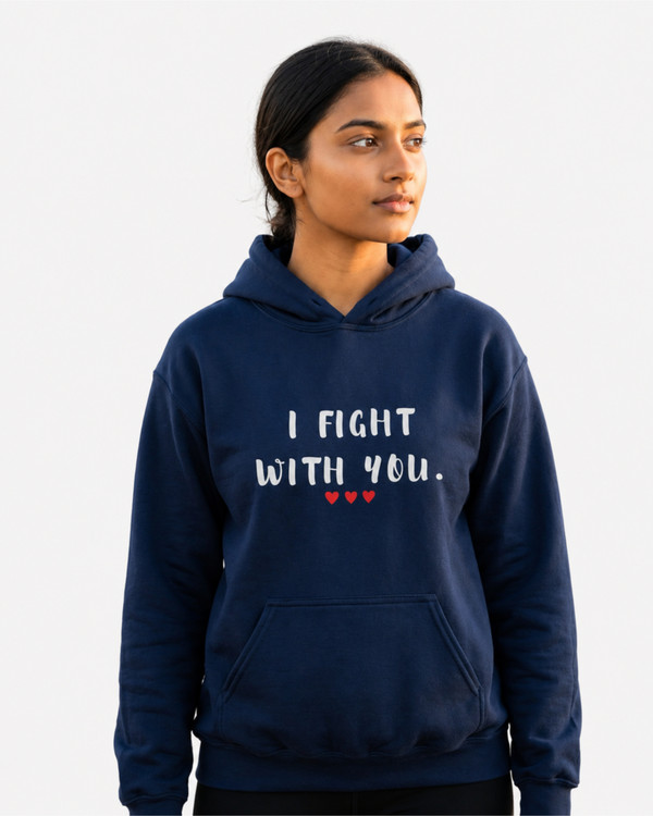 Woman standing on a seaside boardwalk wearing a navy blue pullover hoodie featuring the supportive phrase “I Fight With You” in distressed white lettering with three red hearts beneath. The hoodie has a drawstring hood, front kangaroo pocket, and relaxed fit. The design symbolizes solidarity, encouragement, strength, and unwavering support through life’s challenges.